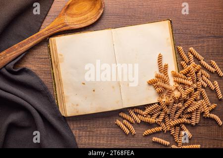 Open blank recipe book and raw whole grain fusilli pasta on the wooden table. Top view. Stock Photo