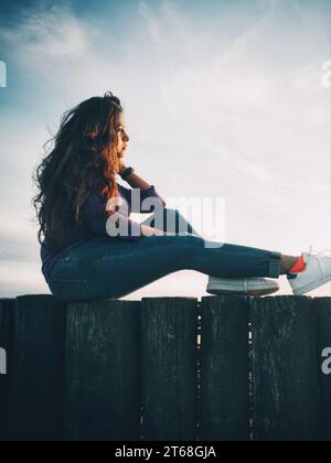 A young woman with long, curly hair sits on a wooden fence at golden hour Stock Photo