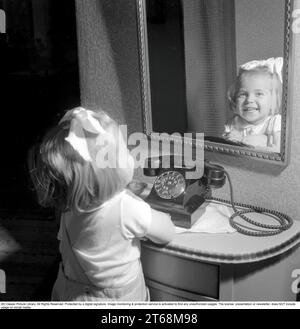 In the 1950s. A little girl pictured by a telephone, smiling and looking a buit guilty as if she was caught playing with the telephone. Sweden 1950. Conard ref 1526 Stock Photo