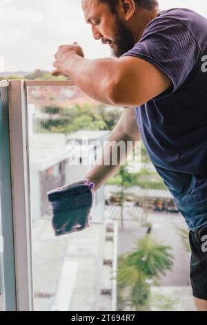 A young man wearing safety glasses and a blue work vest is standing on a ladder, cleaning the window sill with a sponge Stock Photo