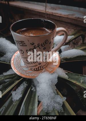 A white porcelain cup of coffee sits atop a saucer on a round plate, surrounded by a layer of freshly fallen snow Stock Photo