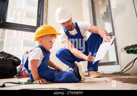 Male builder showing architectural drawings to little boy. Child in work overalls and construction helmet sitting on the floor and studying building plan with father in apartment under renovation. Stock Photo