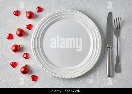 Empty white plate with cutlery on a gray concrete table surrounded by red Christmas balls and hearts. Template for displaying food on the menu. Top vi Stock Photo