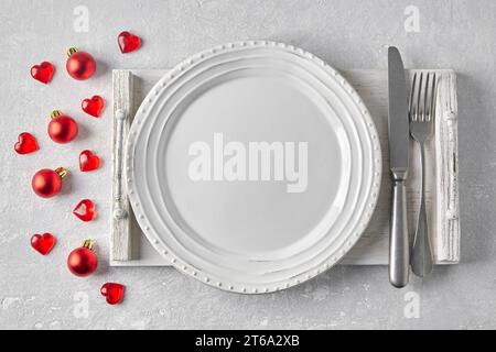 Empty white plate with cutlery on a white wooden tray on a gray concrete table surrounded by red Christmas balls and hearts. Template for presenting f Stock Photo