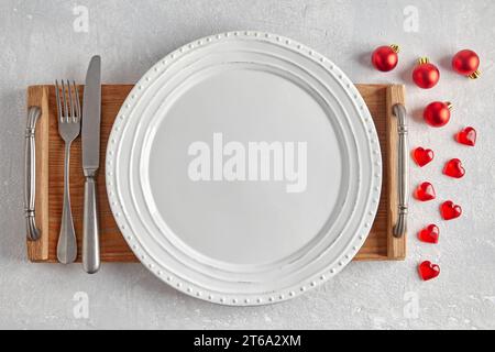 Empty white plate with cutlery on a brown wooden tray on a gray concrete table surrounded by red Christmas tree balls and hearts. Template for present Stock Photo