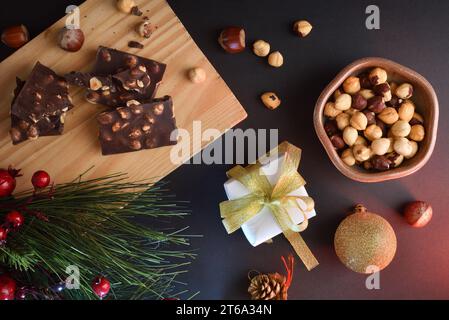 Sweets and with hazelnuts and dried fruit in a bowl on a black table decorated with gift, pine leaves and pine cone at Christmas with lights. Top view Stock Photo