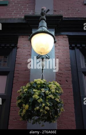An industrial-style street lamp is affixed to the side of an aged brick building, with a decorative planter beneath it Stock Photo