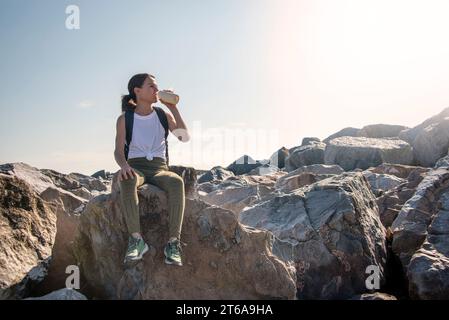 A woman hiker sits on the rock resting in the sun, drinking water from a bottle Stock Photo