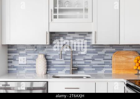 A kitchen faucet detail with a blue glass tile backsplash, white cabinets, decorations on the marble countertop, and a stainless faucet. Stock Photo