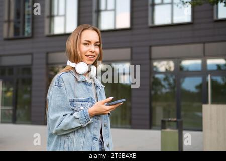 Smiling young woman with headphones on neck looks in camera holding phone. Joyful lady in denim jacket texts best friend after work to hang out Stock Photo