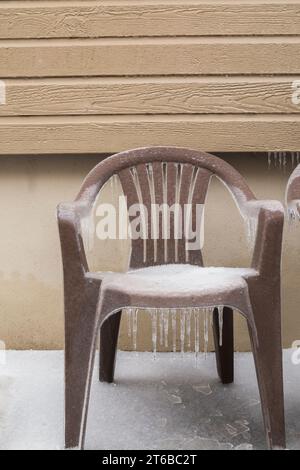 Brown plastic garden armchair covered with ice and snow next to house wall. Stock Photo