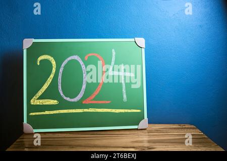 Front view of the number 2024 written in chalk on green chalkboard placed on wooden table against flat blue wall. New year and back to school concept. Stock Photo