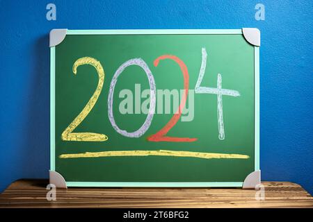 Front view of the number 2024 written in chalk on green chalkboard placed on wooden table against flat blue wall. New year and back to school concept. Stock Photo