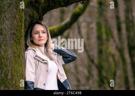 woman in a light-colored coat leaning against a mossy tree Stock Photo
