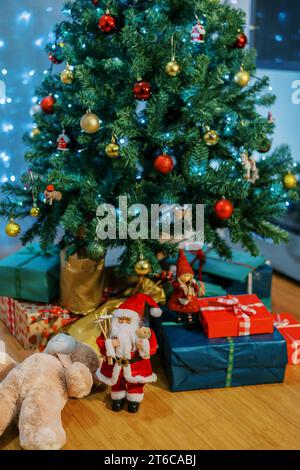 Figurine of Santa Claus stands near a teddy bear and a mountain of gifts under a decorated Christmas tree Stock Photo
