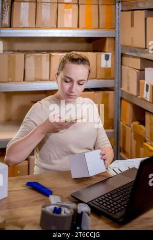 Female small business owner using mobile app on smartphone checking parcel box. Warehouse worker, seller holding phone scanning retail dropshipping Stock Photo