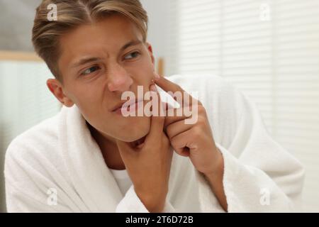 Upset young man looking at mirror and popping pimple on his face indoors. Acne problem Stock Photo