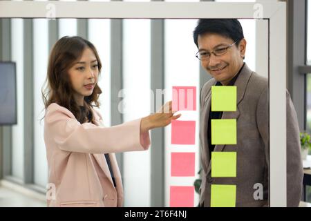 A professional man and woman stand together, engaged in a brainstorming session, using colorful paper notes on a transparent glass wall. Stock Photo