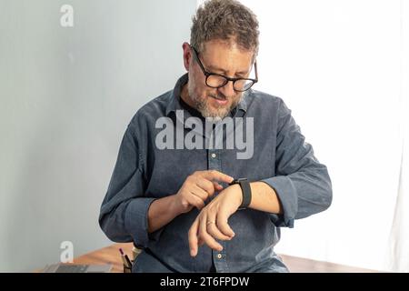A middle-aged man with grey hair and glasses smiling as he interacts with his smartwatch, showcasing the integration of modern wearable technology in Stock Photo