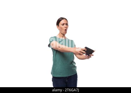 well-groomed young brunette woman dressed in a green t-shirt and jeans plays a game on the phone Stock Photo