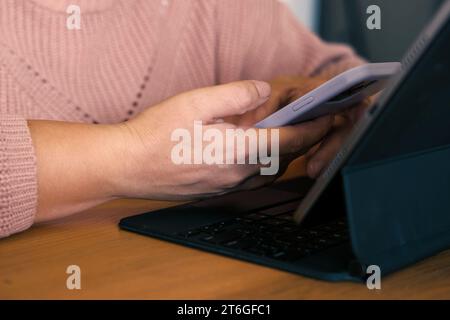 Female using phone looking at laptop or digital tablet device. Unrecognizable woman sitting at table in a cafe and use online banking with smartphone. Stock Photo