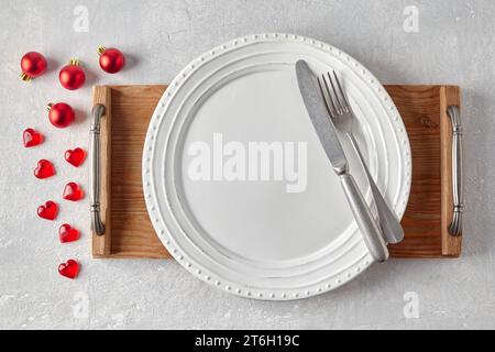 Ceramic white plate with cutlery on a white wooden tray on a gray concrete table surrounded by red Christmas balls and hearts. Layout for displaying f Stock Photo