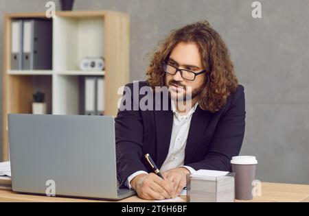 Young businessman sitting at office desk, using laptop computer and taking notes Stock Photo