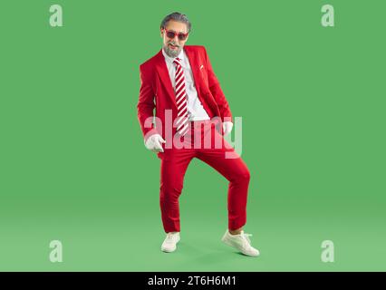Senior grey haired showman in red sunglasses and red suit, white gloves posing on green background. Stock Photo