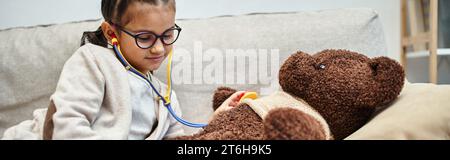 happy kid in casual wear and eyeglasses playing doctor with teddy bear on sofa in living room Stock Photo
