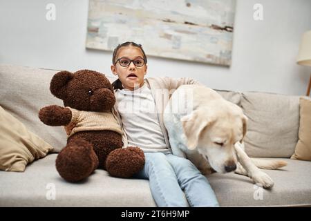 scared girl in eyeglasses holding teddy bear and sitting on sofa with labrador while watching movie Stock Photo