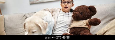 scared girl in eyeglasses holding teddy bear and sitting with labrador while watching movie, banner Stock Photo