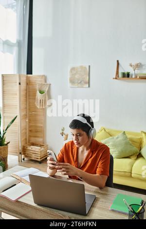 vertical shot of young student studying at laptop with phone in hands with living room on backdrop Stock Photo