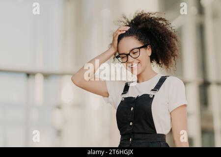Laughing young woman adjusting her glasses, enjoying a sunny day in a chic denim jumper Stock Photo