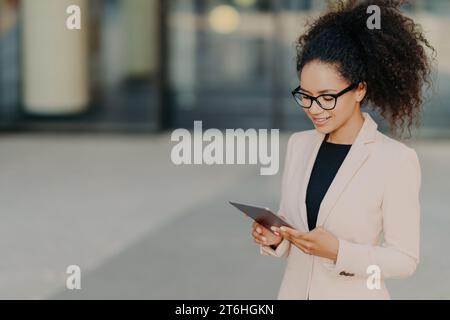 Focused professional woman using a tablet, in a stylish blazer and glasses, urban backdrop. Stock Photo