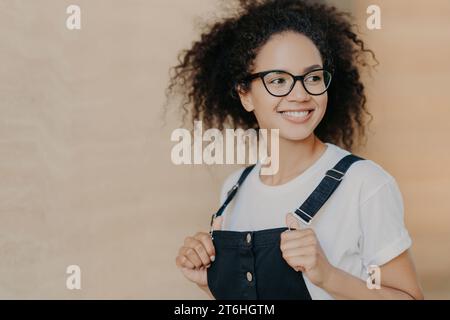 Optimistic young lady with curly hair adjusting her overalls, looking away with a smile Stock Photo