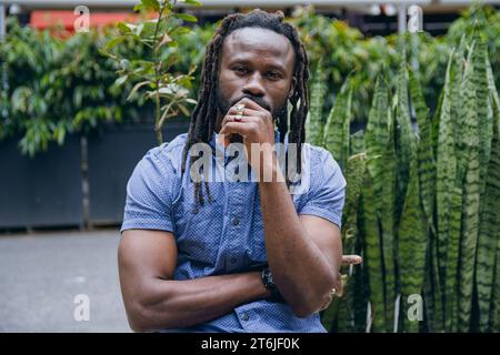 Strong handsome young black man with dreadlocks, beard and shirt, sitting outdoors thoughtfully with hand on lips and looking at camera, front view, p Stock Photo