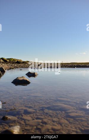 A rock pool at low tide on a clear sunny day, with the bottom visible through the calm water and rocks reflecting on it's surface. Stock Photo