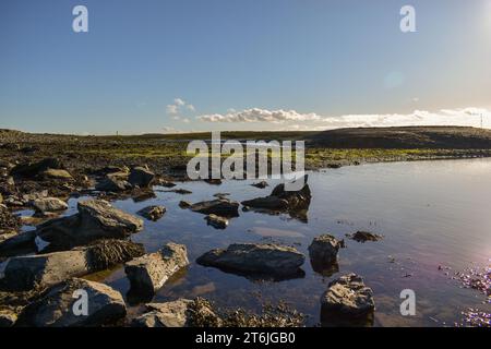 A large rock pool at low tide on a clear sunny day, surrounded by seaweed and algae with some rocks reflecting on the surface of the water. Stock Photo