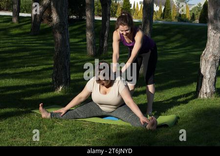 Yoga trainer conducts personal training for overweight woman in nature. fat woman is doing Pilates in city park under guidance of instructor. Healthy Stock Photo