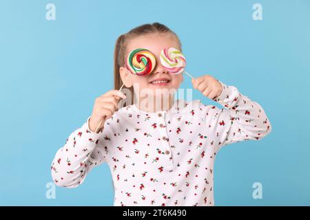 Happy little girl covering eyes with colorful lollipops on light blue background Stock Photo