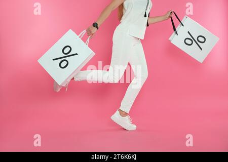 Discount, sale, offer. Woman running with shopping bags against pink background, closeup. Paper bags with percent signs Stock Photo