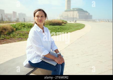 Attractive young woman sitting on the bench with laptop, relaxing during lunch time. Pensive business woman thoughtfully looking to the distance, dres Stock Photo