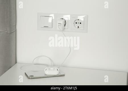 Charging case of modern wireless earphones plugged into power socket and smartphone on white table indoors Stock Photo