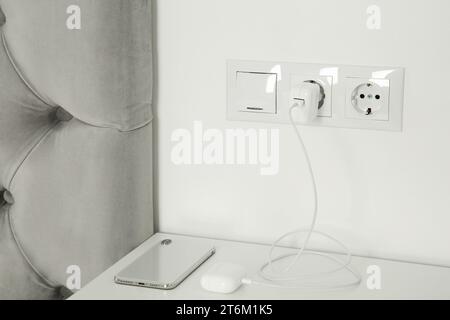 Charging case of modern wireless earphones plugged into power socket and smartphone on white table indoors Stock Photo