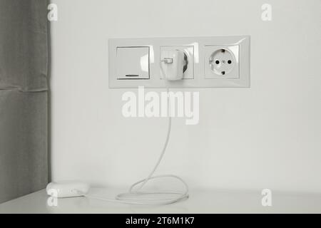 Charging case of modern wireless earphones plugged into power socket on white table indoors Stock Photo