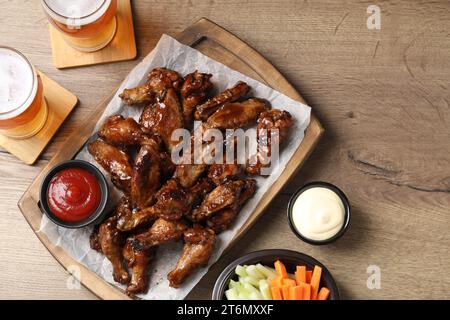 Tasty chicken wings, glasses of beer, sauces and vegetable sticks on wooden table, flat lay. Delicious snacks Stock Photo