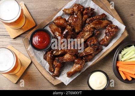 Tasty chicken wings, glasses of beer, sauces and vegetable sticks on wooden table, flat lay. Delicious snacks Stock Photo