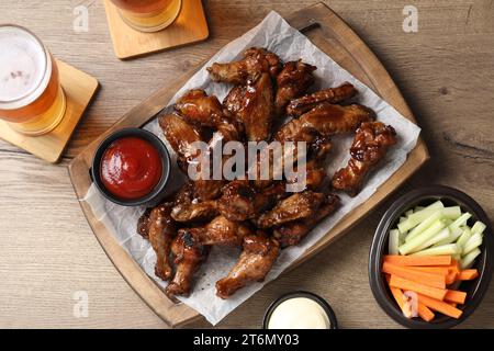 Tasty chicken wings, glasses of beer, sauces and vegetable sticks on wooden table, flat lay. Delicious snacks Stock Photo