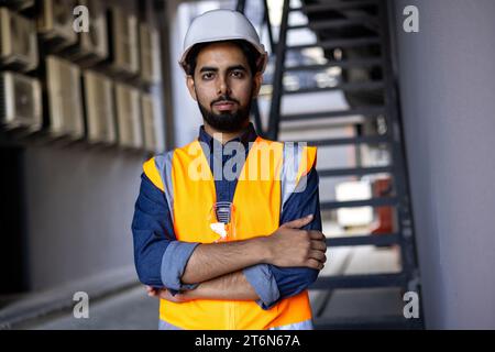 Serious engineer construction worker in helmet standing near building with crossed arms, arab work foreman looking and camera, senior project worker designer. Stock Photo