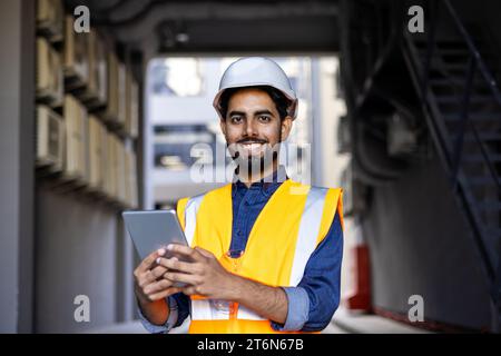 Happy engineer builder in helmet standing near building with tablet in hands, arab work foreman looking and smiling at camera, senior project worker designer leader concept. Stock Photo